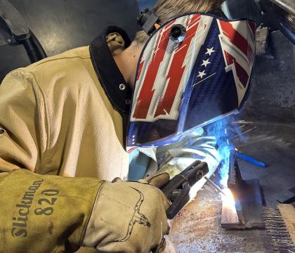 Student welding in a shop with a USA graphic welding helmet.